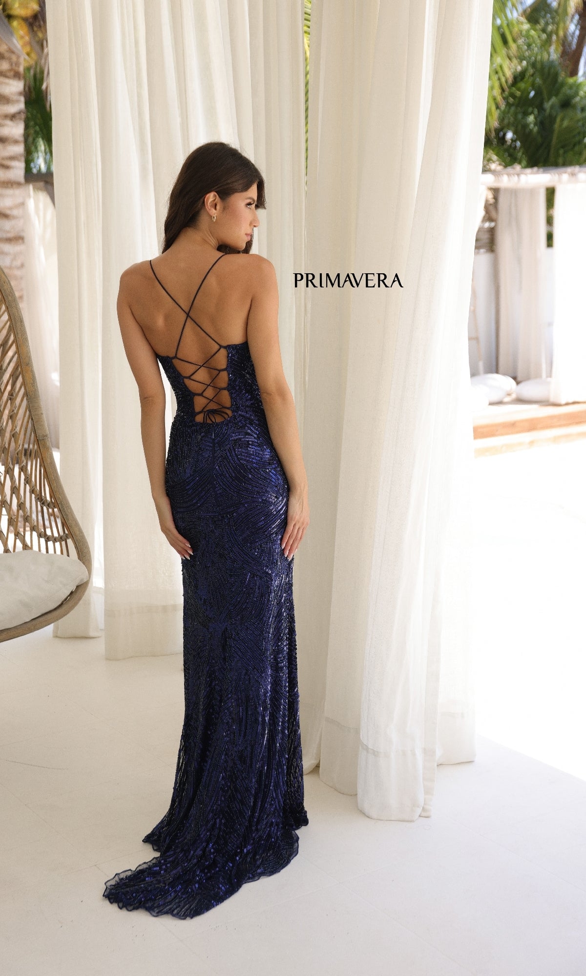A woman stands near white curtains in a bright outdoor setting, wearing the Strappy-Back Long Sequin Prom Gown: Primavera 4365 in navy blue with crisscross straps.